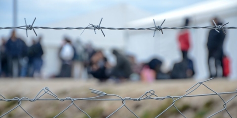 Line of people off focus seen through bar-wired fence